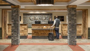 A reception desk with a stone wall backdrop and wooden accents, a person with a suitcase, and a television screen above the desk. - Killington Grand Hotel (Killington)