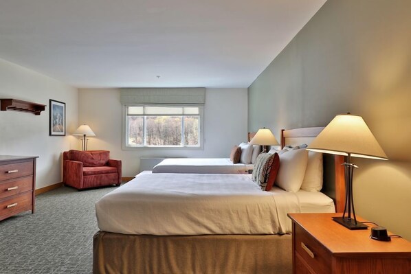 A hotel room with two beds, a desk, a chair, a lamp, and a window with a view of trees.