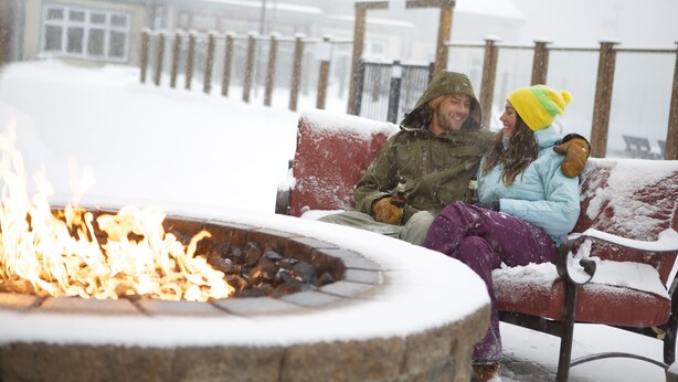 A bench with snow accumulation, a fire pit with visible flames, and a wooden fence in the background. - Killington Grand Hotel (Killington)