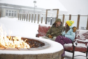 A bench with snow accumulation, a fire pit with visible flames, and a wooden fence in the background. - Killington Grand Hotel (Killington)