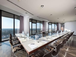A conference room with a long table, chairs, and large windows offering a city view.