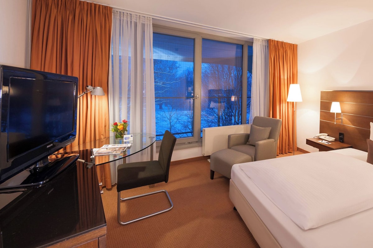 Et hotelværelse med en seng, et skrivebord med fjernsyn, en stol og et natbord med lampe.