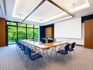 A conference room with a long table, blue chairs, a projector screen, and a view of greenery through large windows.