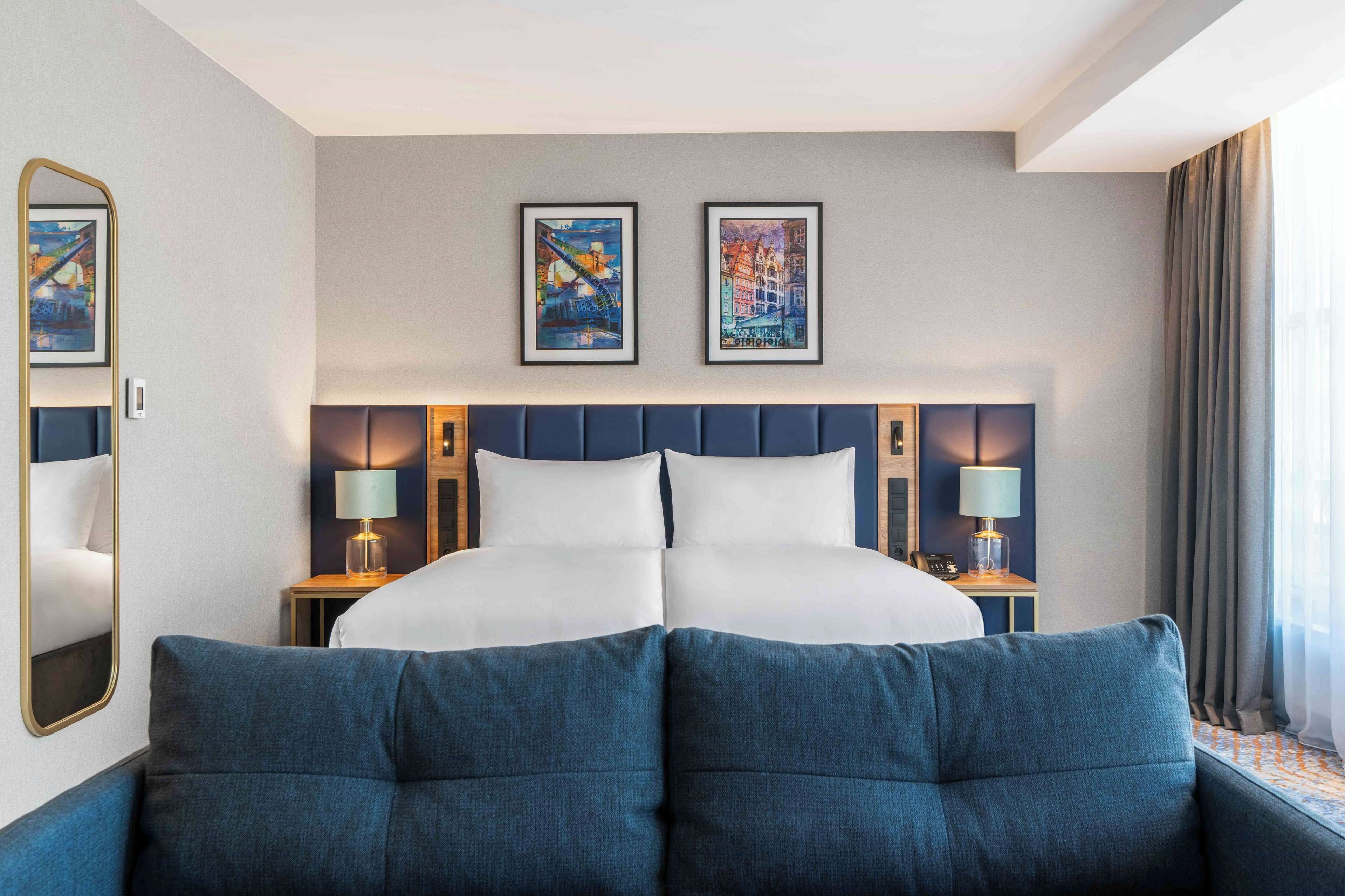 Habitació d'hotel amb un llit gran, dues taules de nit amb làmpades, un capçal de llit amb obra d'art i una finestra gran amb cortines.