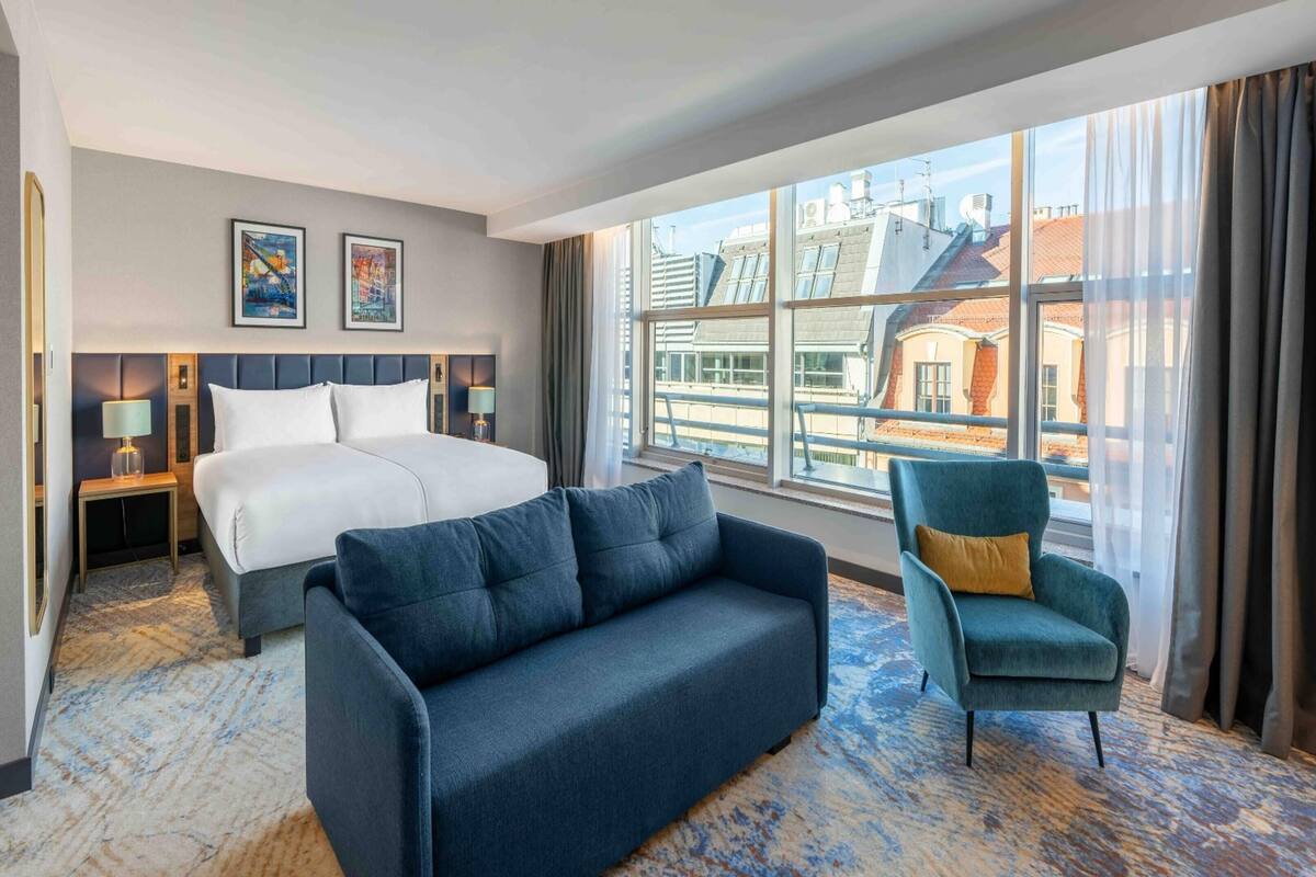 A hotel room with a large bed, a sofa, a chair, and a large window with a view of buildings.