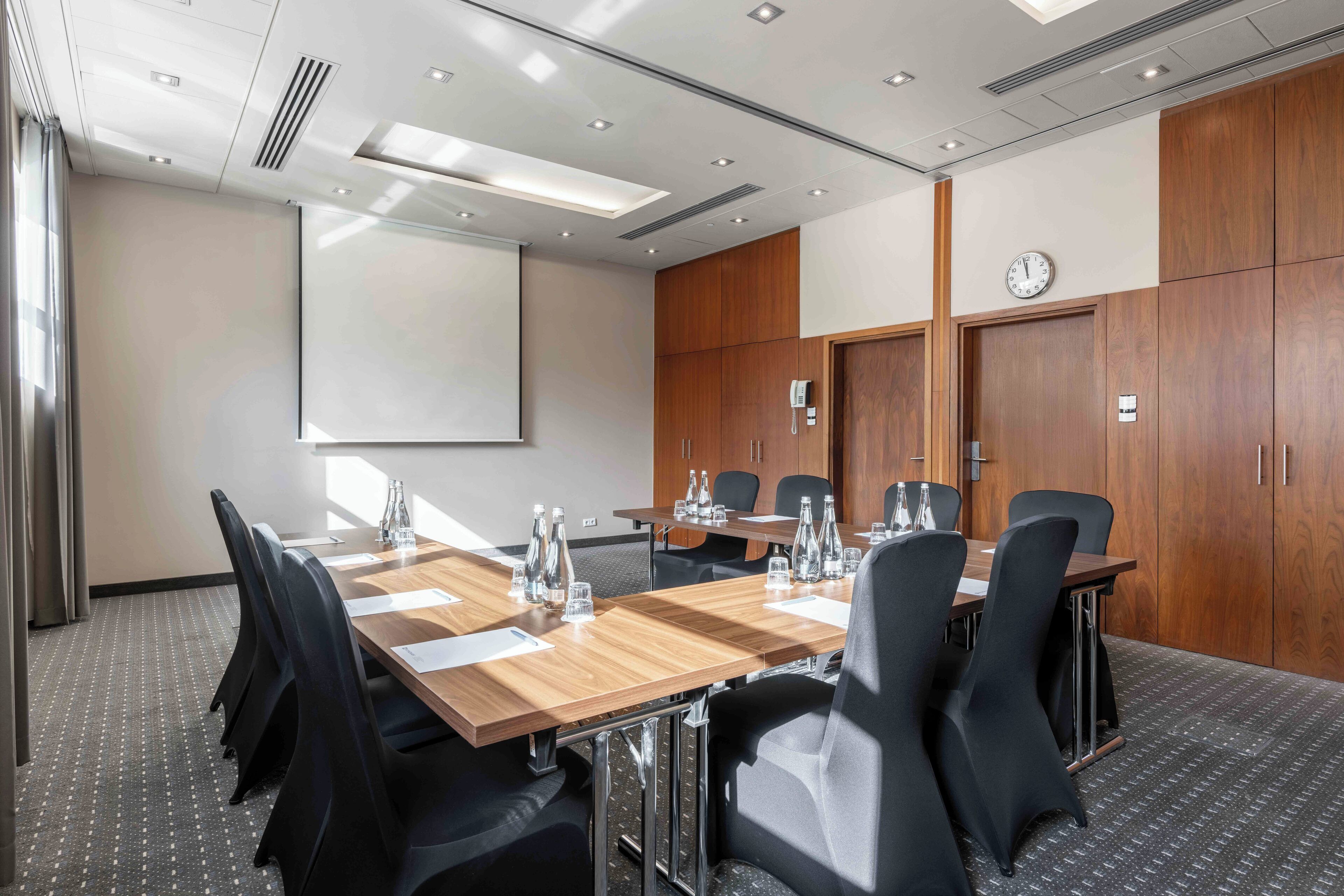 A conference room with a long table, black chairs, a projector screen, wooden cabinets, and a clock on the wall.