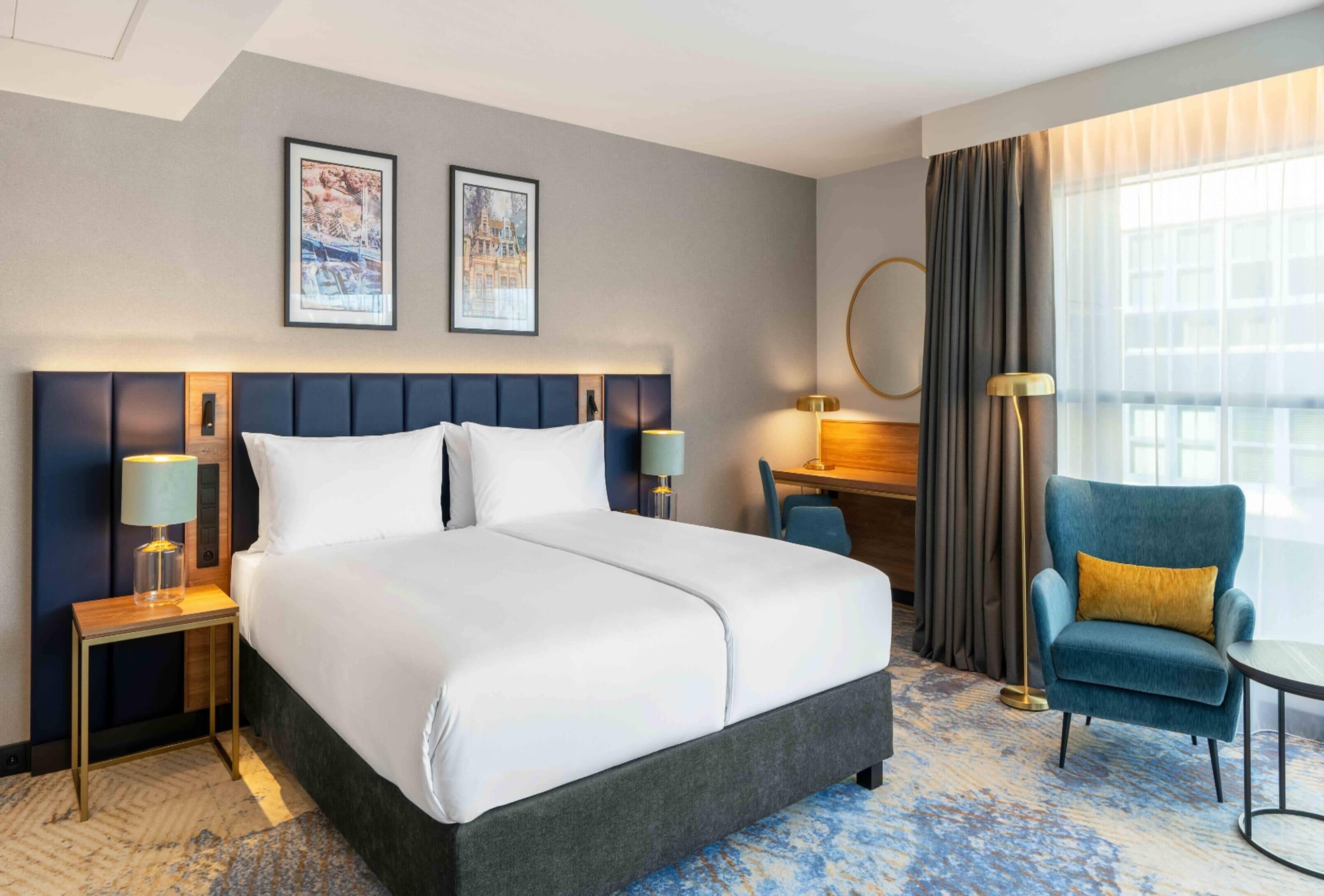 A hotel room with a large bed, two bedside tables with lamps, a desk, a chair, and a large window with curtains.