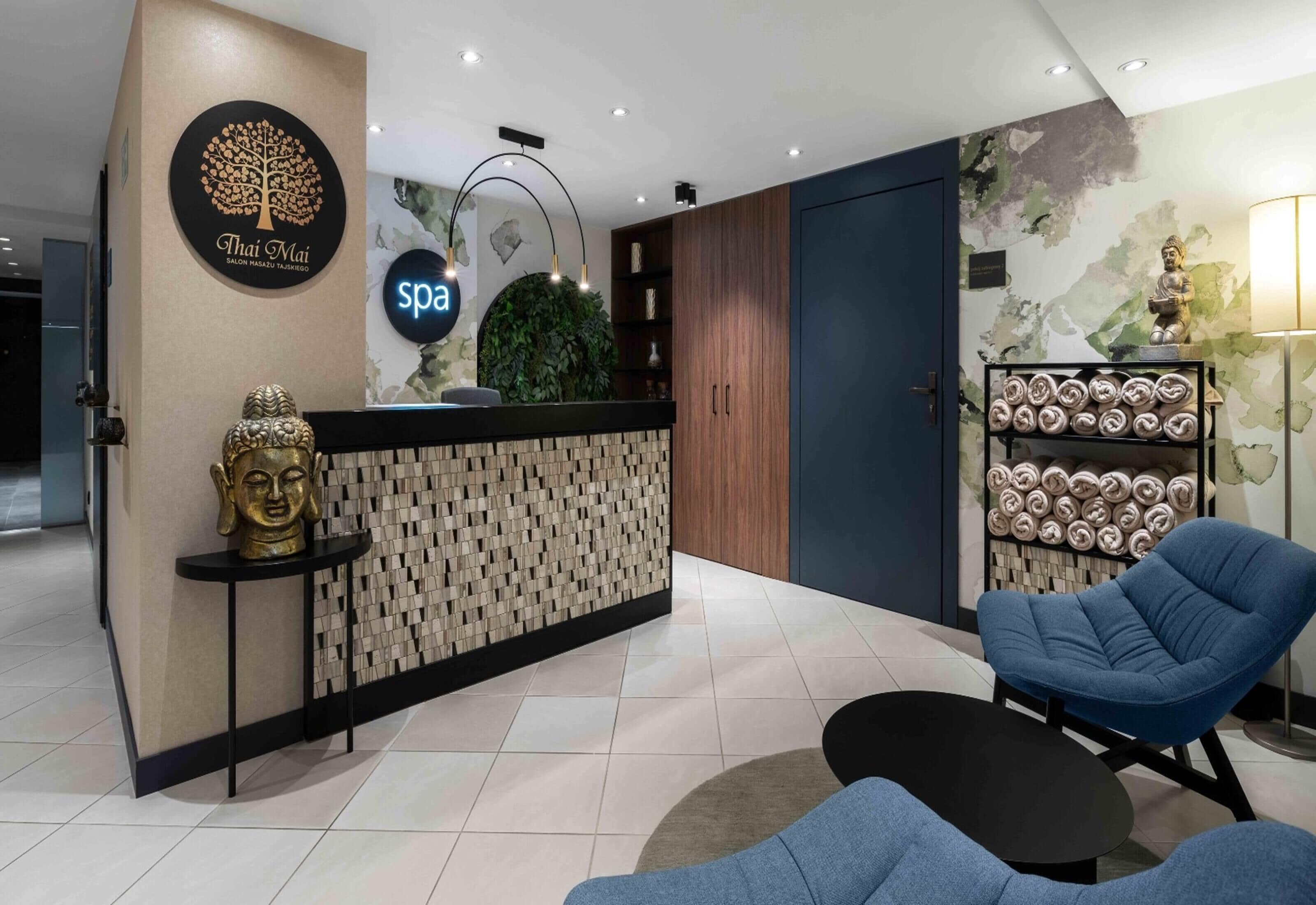 A modern spa reception area with a wooden reception desk, a Buddha statue, a blue chair, and a wall-mounted spa sign.