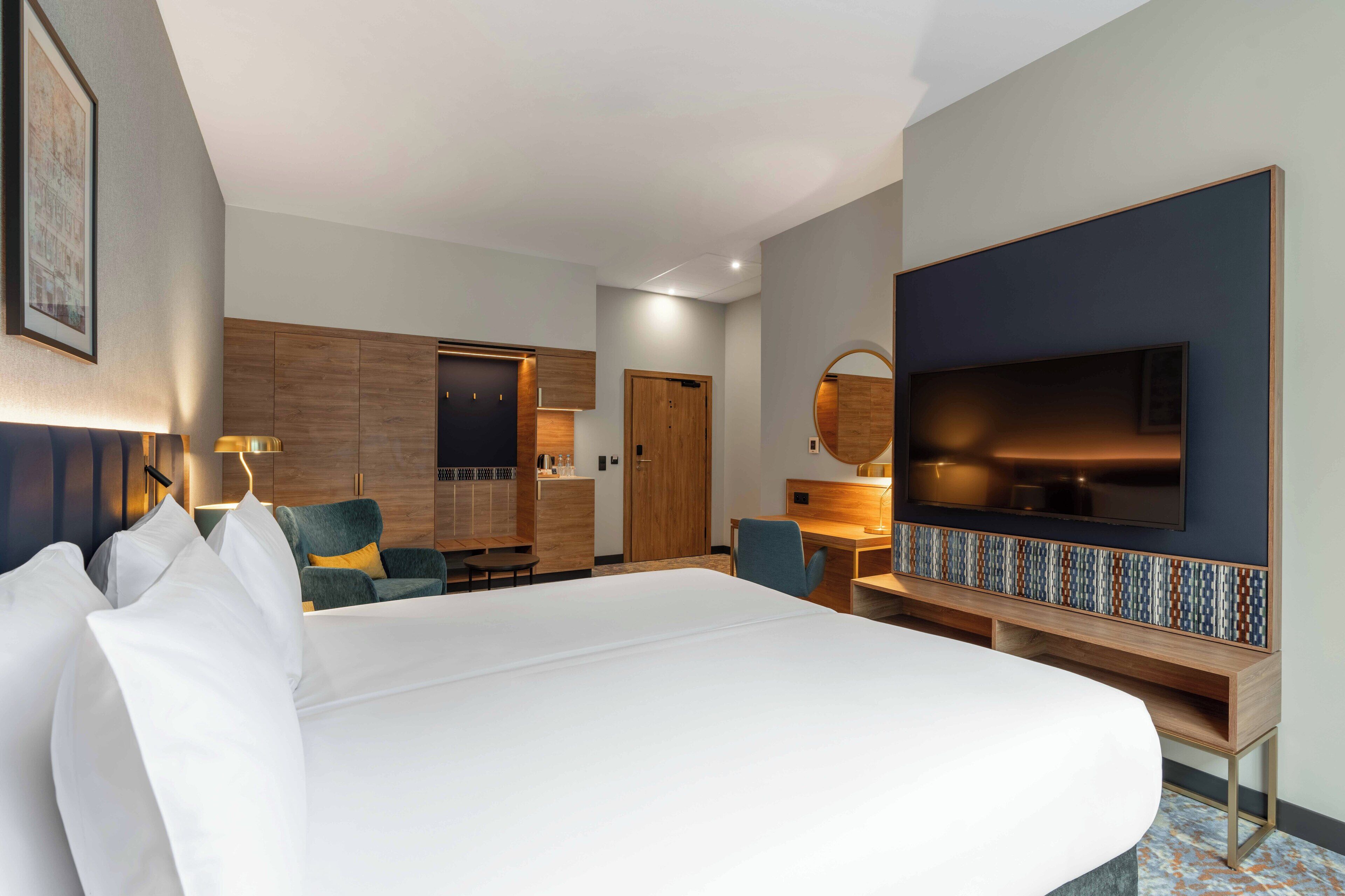 A hotel room with a large bed, a desk, a chair, a TV, and a wooden wardrobe.