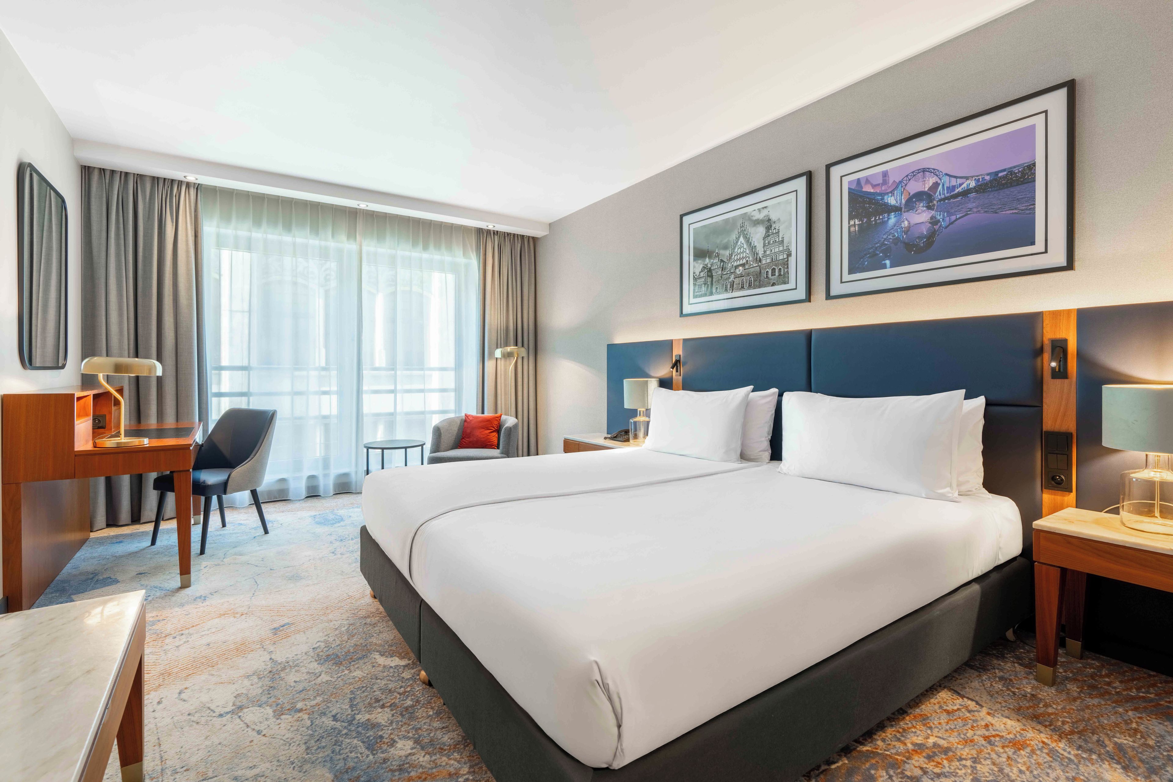 A hotel room with a large bed, a desk, a chair, a nightstand, and two framed pictures on the wall.