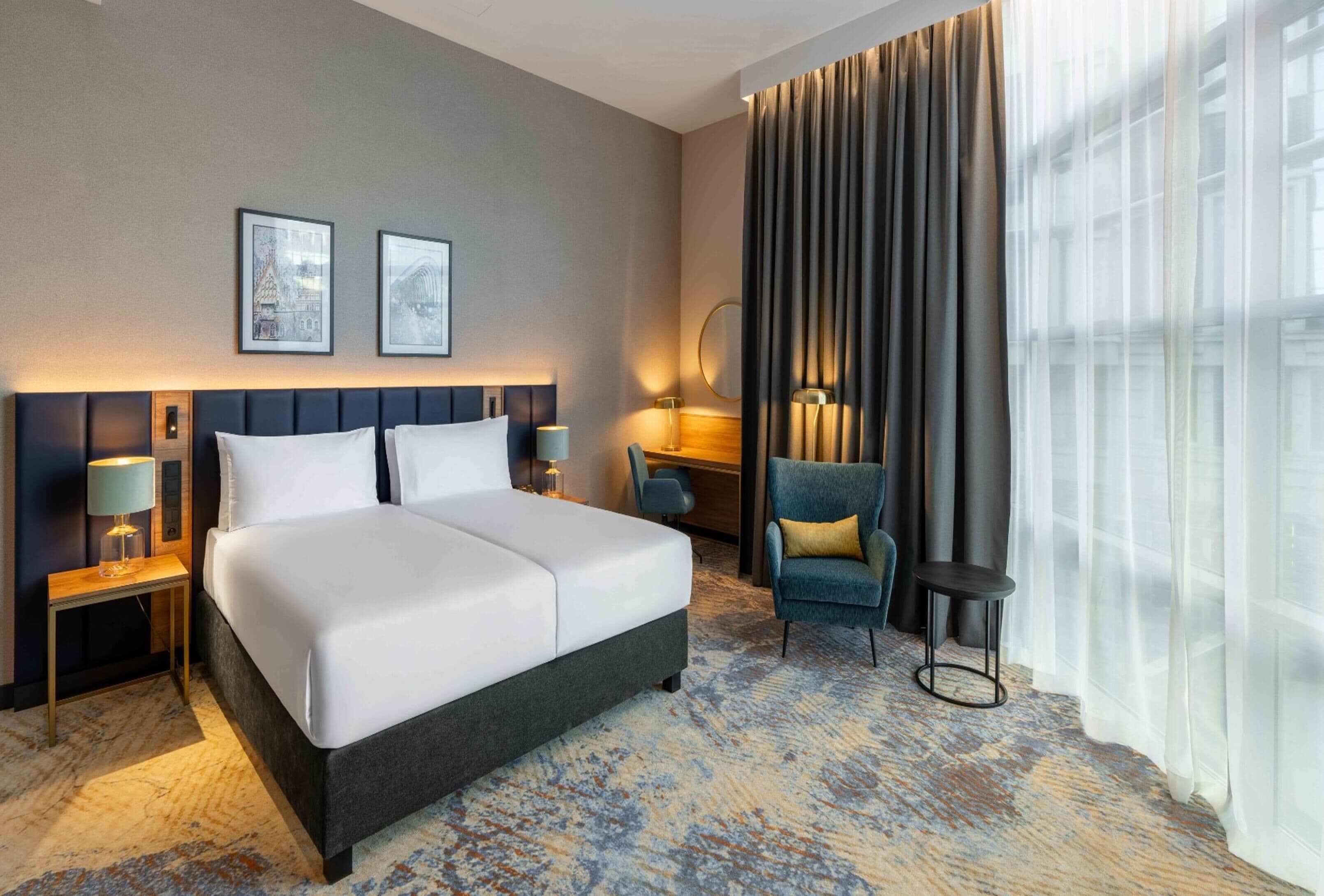 A hotel room with a large bed, two bedside tables with lamps, a chair, a desk, and a large window with curtains.
