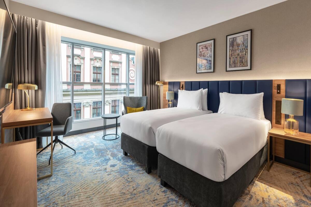 A hotel room with two beds, a desk, a chair, a window with a view of buildings, and two framed pictures on the wall.