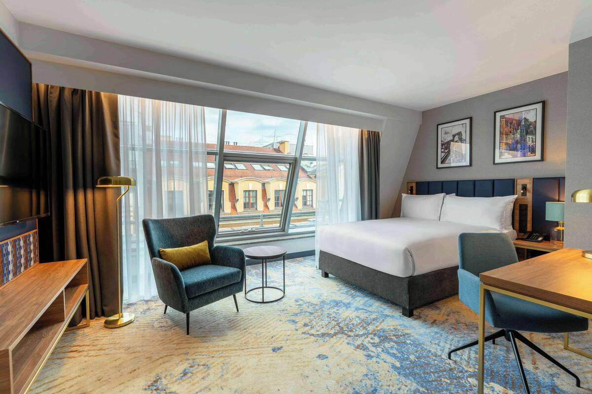 A hotel room with a large bed, a desk, a chair, a TV, and a view of buildings through the window.