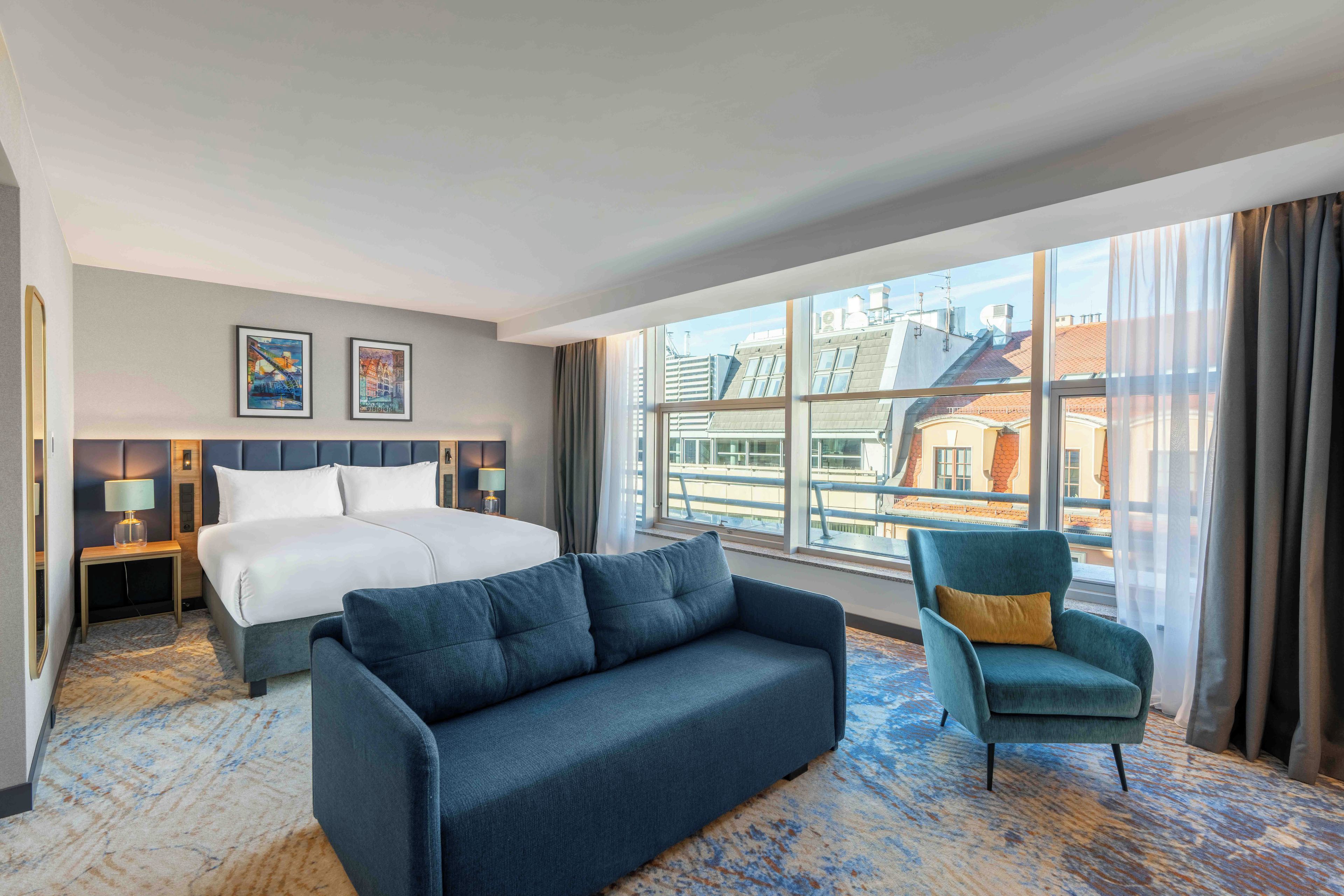 A hotel room with a bed, a sofa, a chair, a nightstand, a lamp, and a large window with a view of buildings.