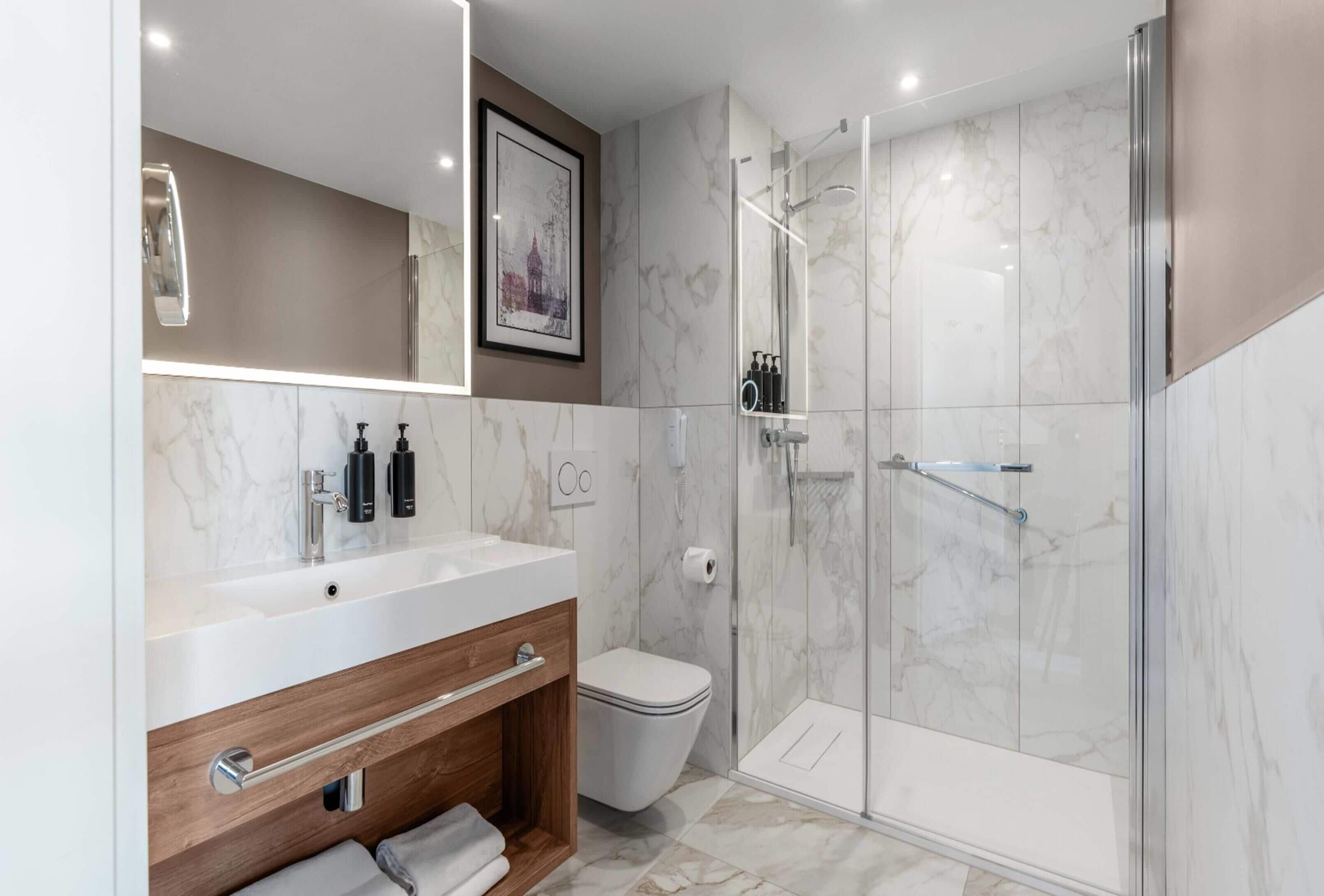 Un baño moderno con lavamanos de mármol, tocador de madera, inodoro y ducha con mampara de vidrio.