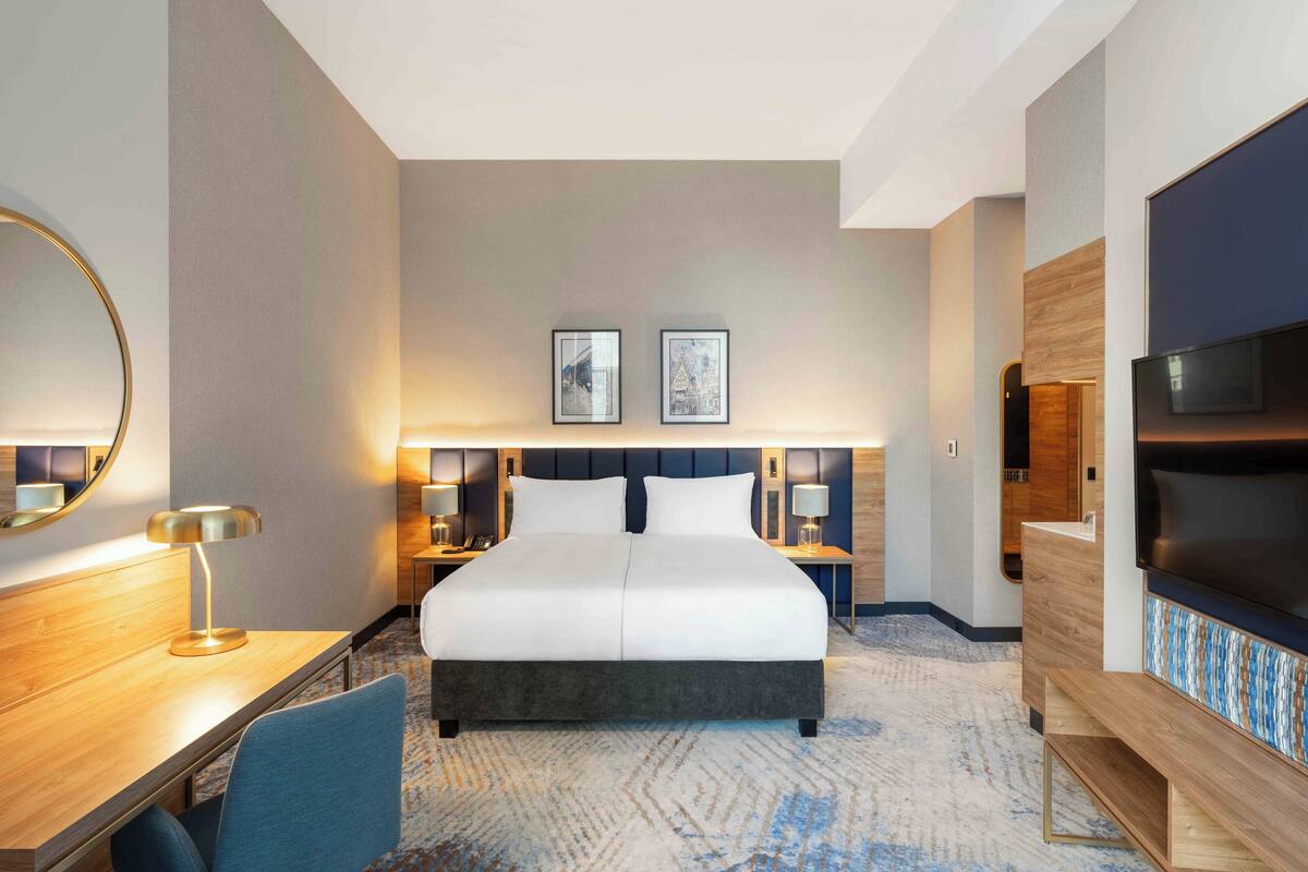 A hotel room with a large bed, bedside tables, a desk, a chair, a TV, and two framed pictures on the wall.