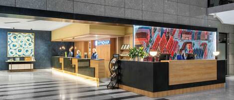 A hotel reception area with a modern design, featuring a reception desk, a large mural, and a clean, tiled floor.