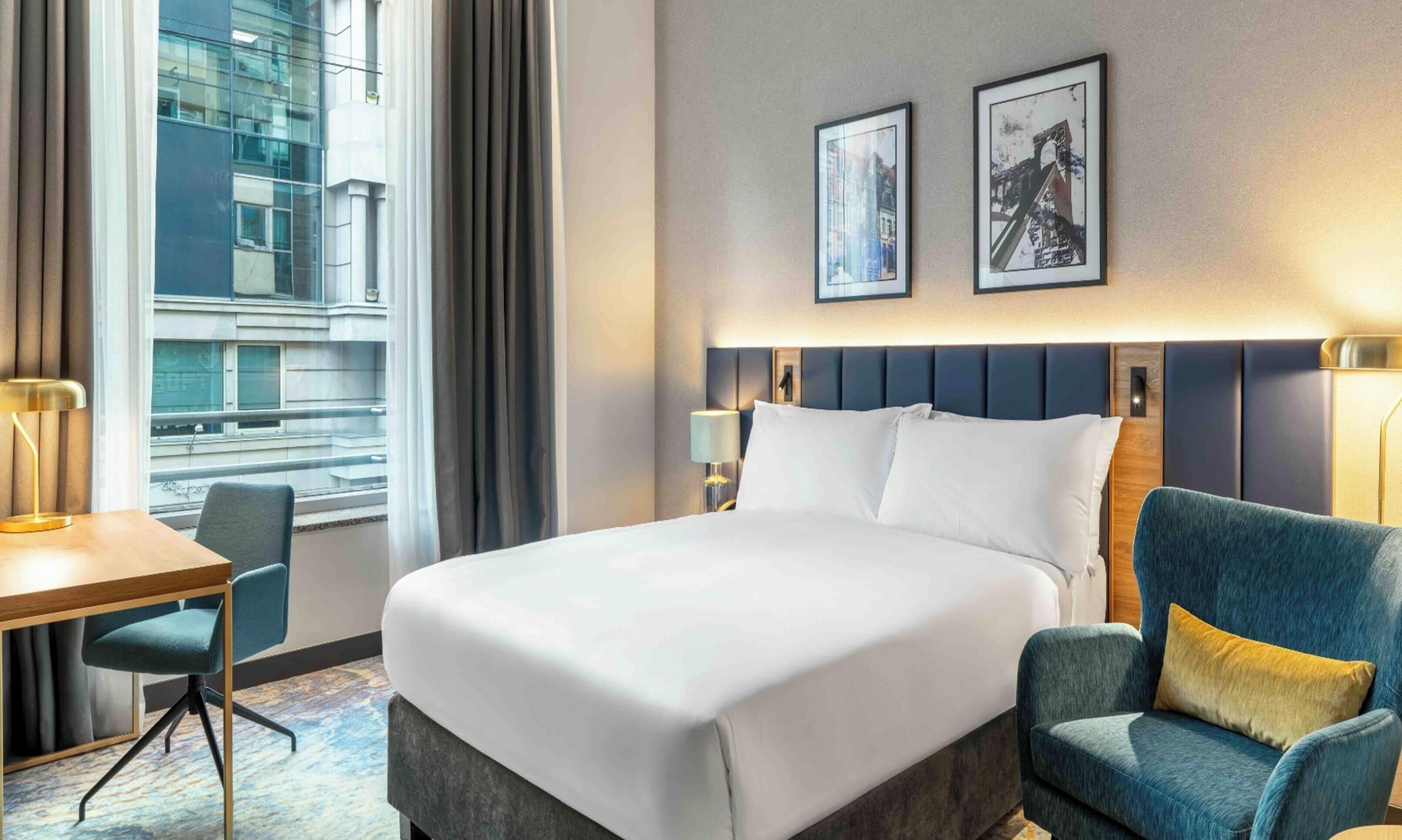 Ein Hotelzimmer mit einem großen Bett, einem Schreibtisch, einem Sessel und Blick auf eine Stadtlandschaft durch das Fenster.