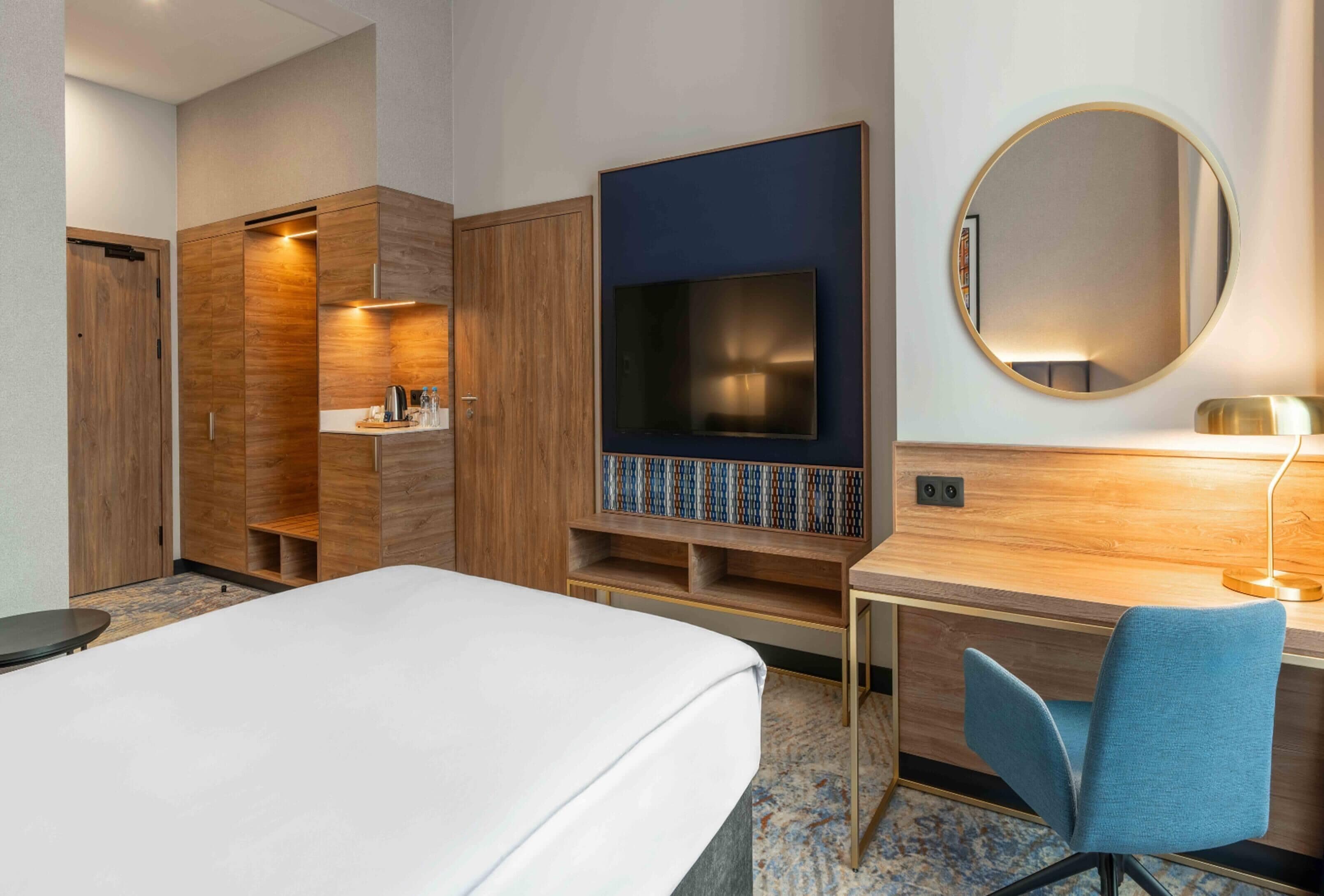 Quarto de hotel com cama, escrivaninha, cadeira, televisão e espelho.