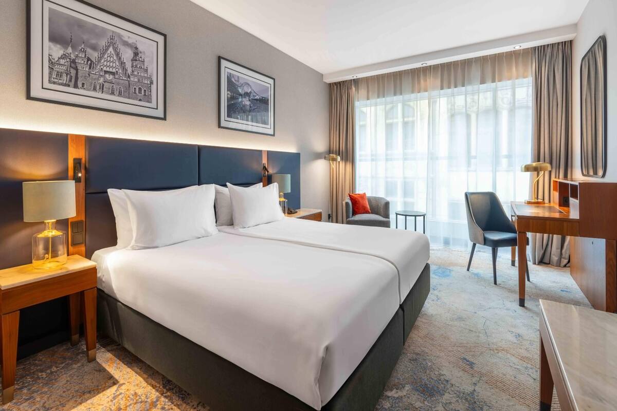 A hotel room with a large bed, a desk, a chair, and two framed pictures on the wall.