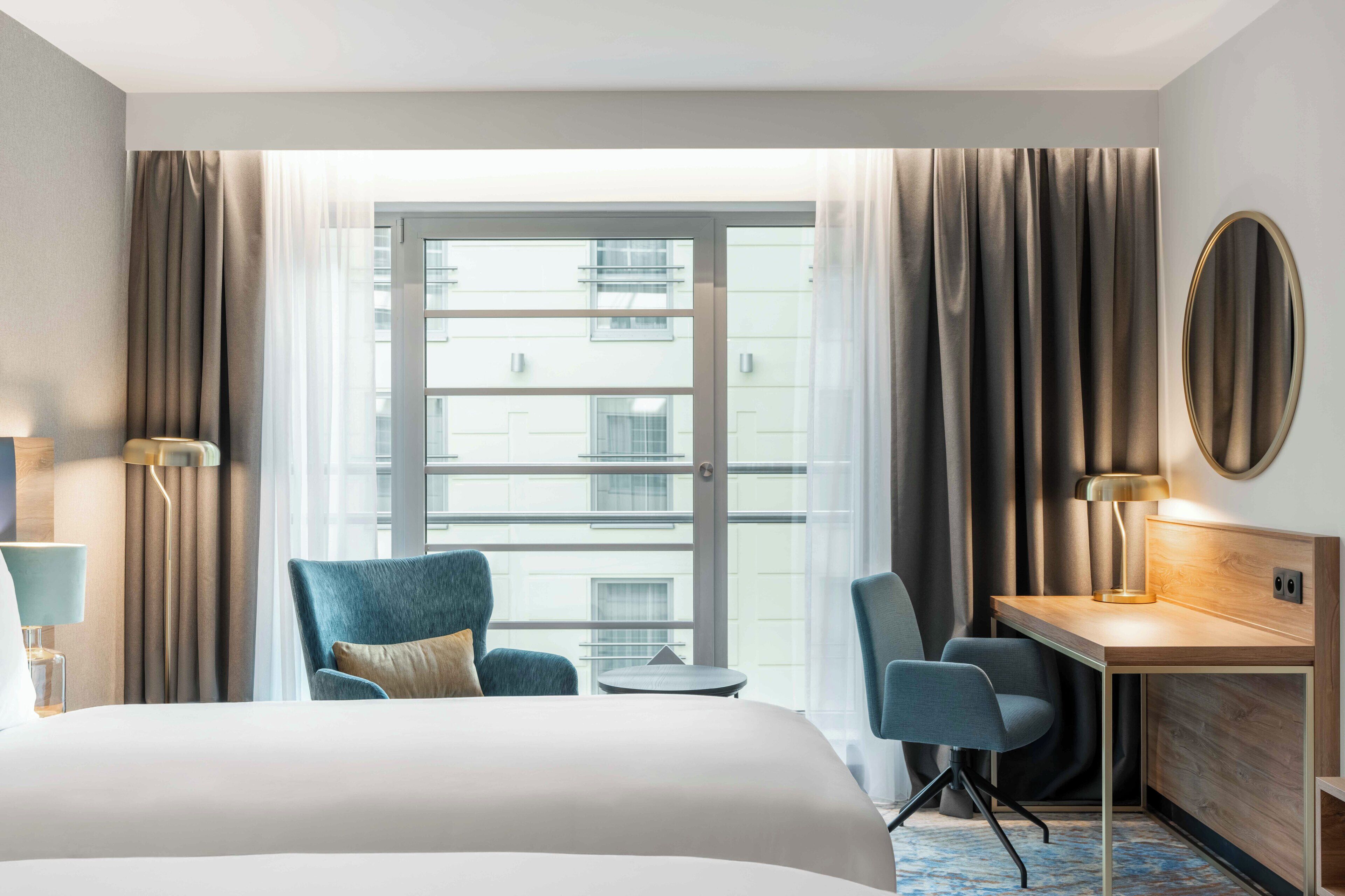 A hotel room with a large bed, a desk, two chairs, a small table, and a large window with curtains.