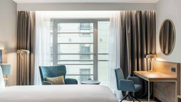 Ein Hotelzimmer mit einem großen Bett, einem Schreibtisch, zwei Stühlen, einem kleinen Tisch und einem großen Fenster mit Vorhängen.