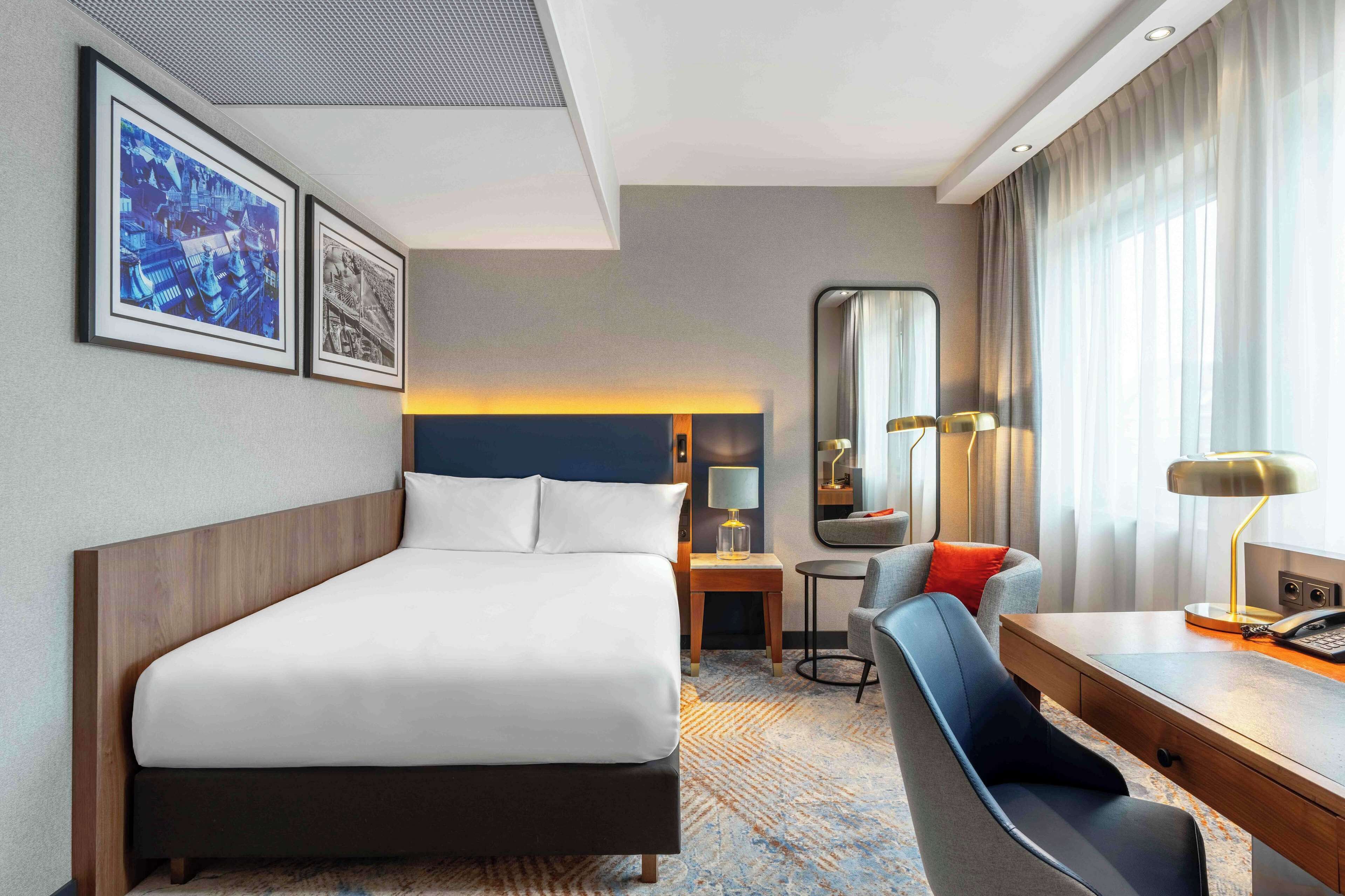 Ein Hotelzimmer mit einem Bett, einem Schreibtisch, einem Stuhl, einem Spiegel und zwei gerahmten Bildern an der Wand.