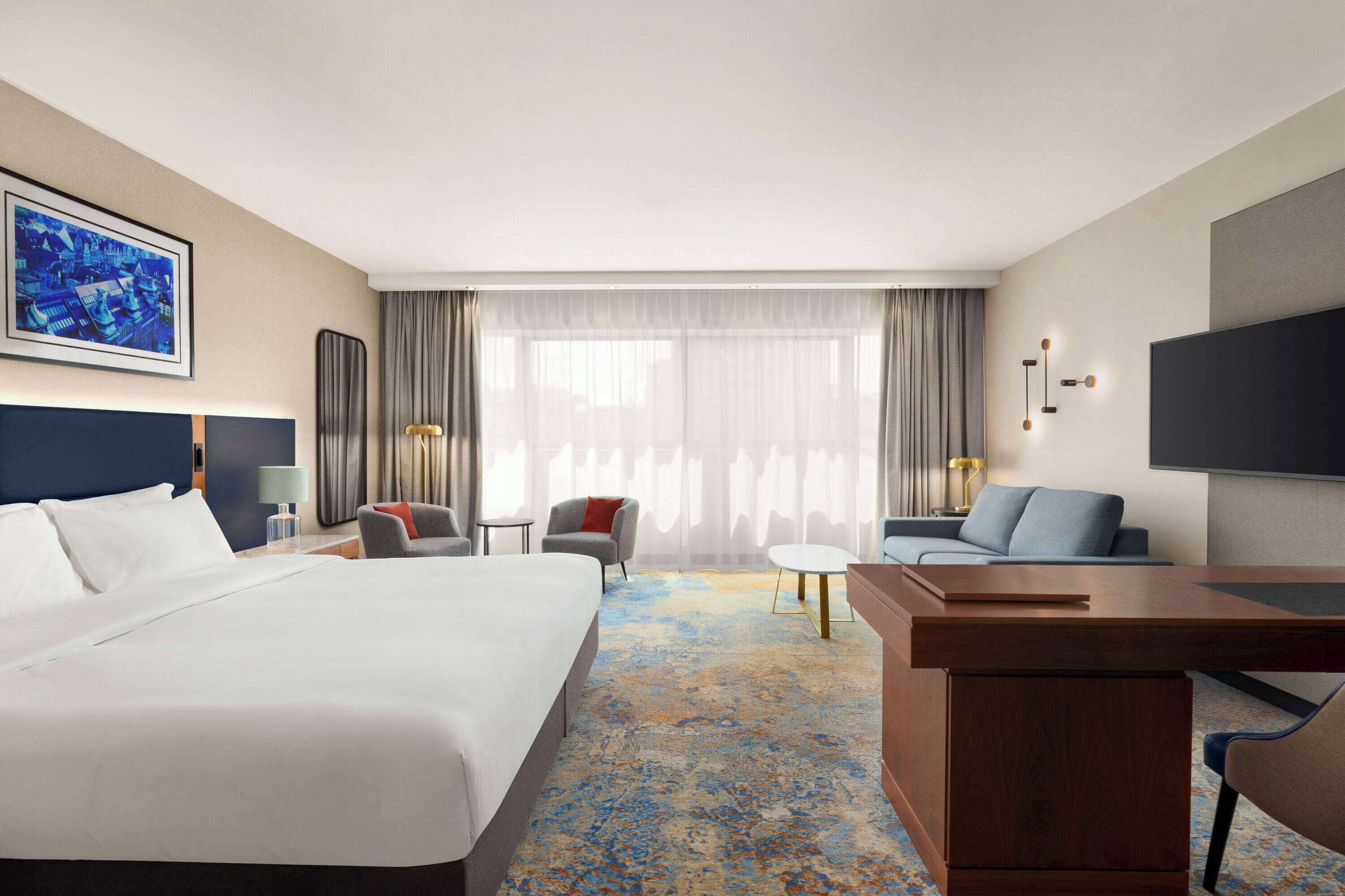 A hotel room with a large bed, a desk, two chairs, a small table, and a TV.