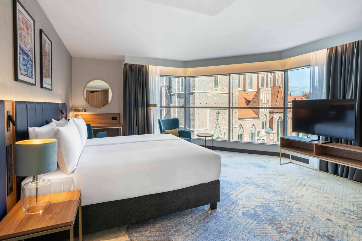 A modern hotel room with a large bed, a TV, a desk, and a view of the city through large windows.
