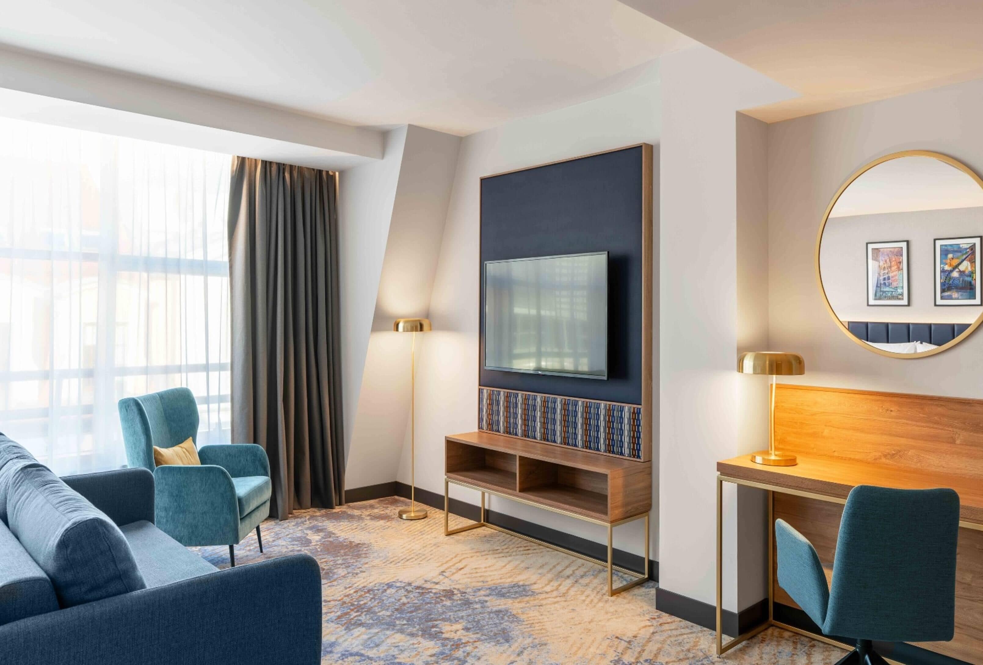 A modern hotel room with a large flat-screen TV mounted on a dark wall, a wooden desk with a lamp, a comfortable sofa, and a round mirror with artwork.