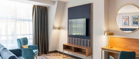 A modern hotel room with a large flat-screen TV mounted on a dark wall, a wooden desk with a lamp, a comfortable sofa, and a round mirror with artwork.