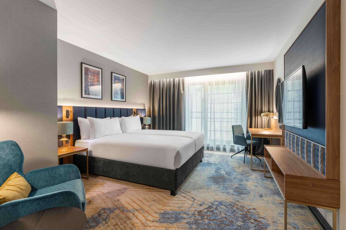 A hotel room with a large bed, a desk, a chair, a TV, and a window with curtains.