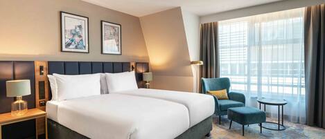 Habitación de hotel con una cama grande, dos mesitas de noche, una silla, una mesita pequeña y un ventanal con cortinas.