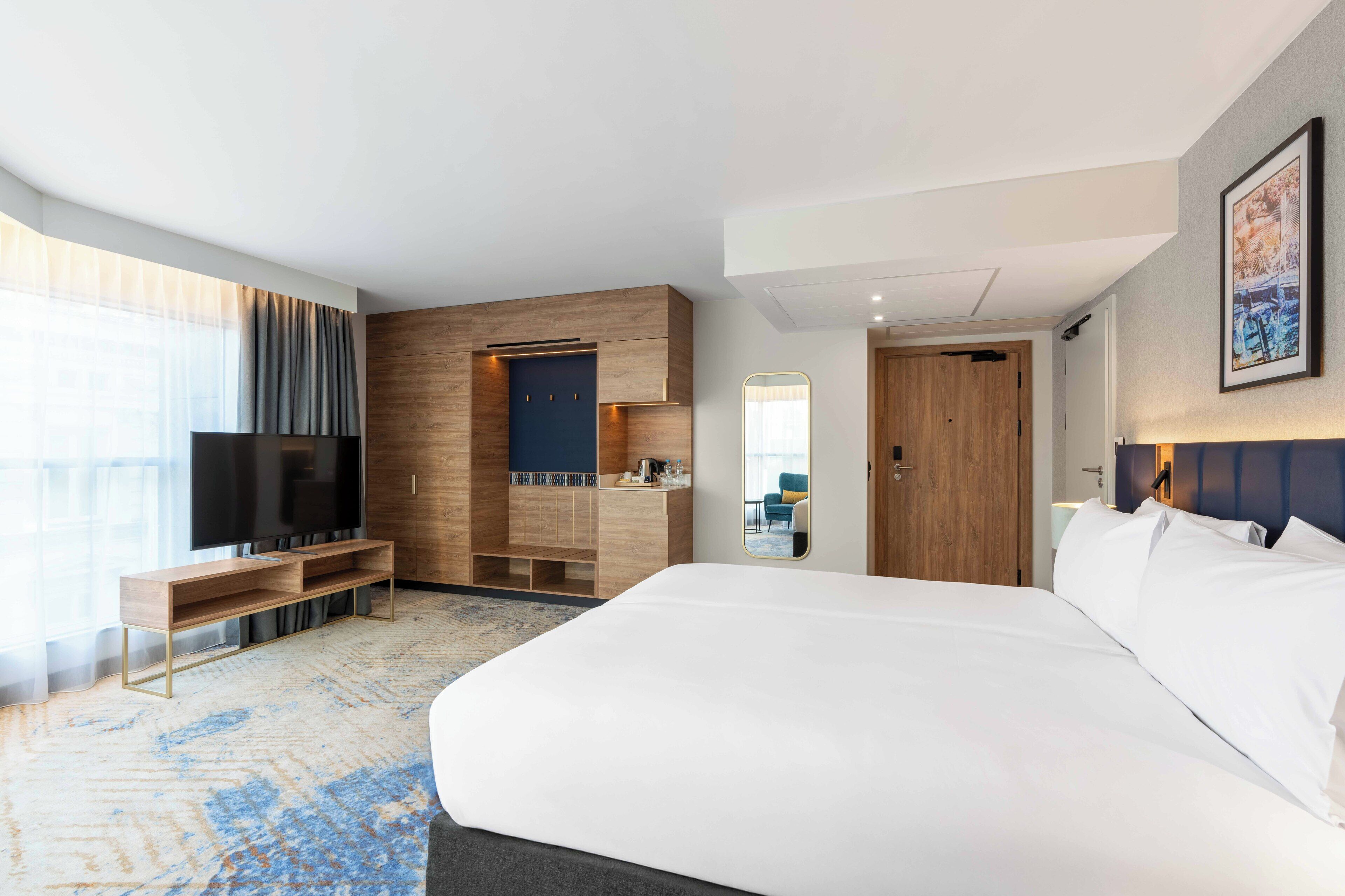A modern hotel room with a large bed, a TV, a wooden wardrobe, and a bathroom visible through a door.