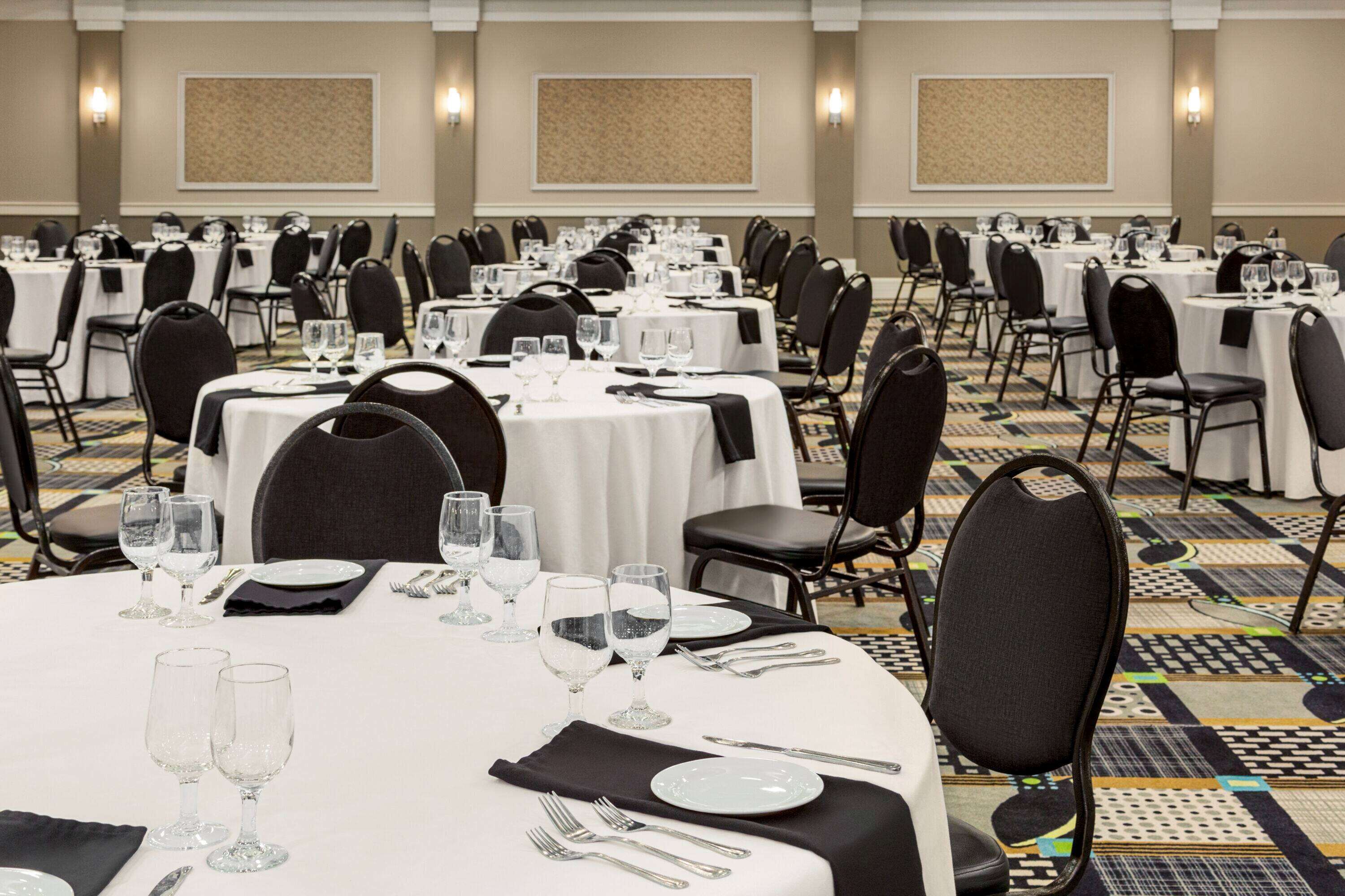 Un salón de banquetes con varias mesas redondas, cada una cubierta con manteles blancos, sillas negras y cubertería.