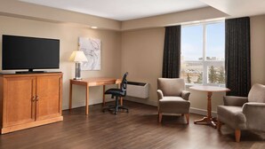 Habitación de hotel con un armario de madera, un televisor de pantalla plana, un escritorio con una silla, una mesita redonda pequeña con dos sillas y un ventanal con cortinas.