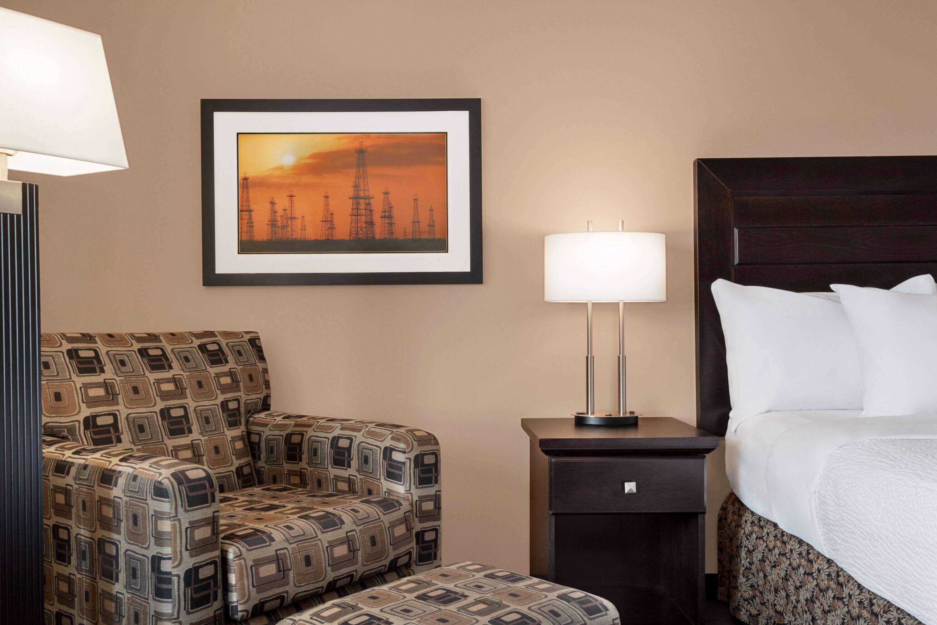A hotel room with a patterned sofa, a bedside table with a lamp, a framed picture on the wall, and a bed with white bedding.