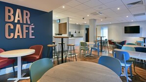 Un bar café moderno con mesas redondas y sillas de colores, una zona de cocina y un televisor de pantalla plana en la pared.
