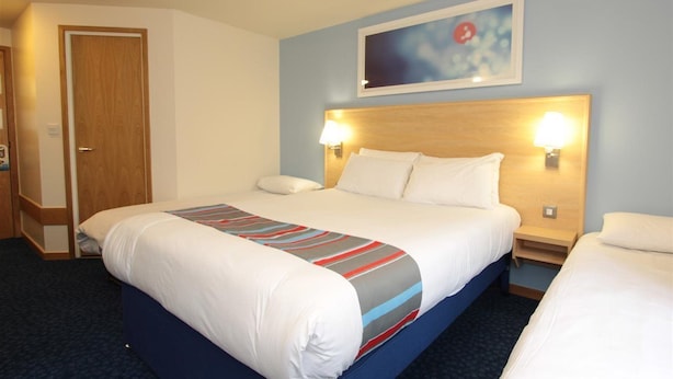 Ein Hotelzimmer mit einem großen Bett, zwei Nachttischen, einer Kopfteil mit Wandlampen, einer Tür und einem gerahmten Bild an der Wand.