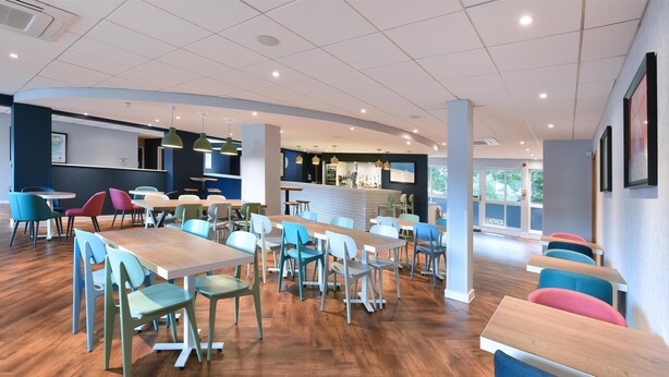 A modern dining area with wooden tables and colorful chairs, a bar in the background, and large windows allowing natural light. - Travelodge Ashford (Ashford)