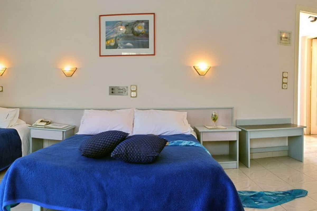 A hotel room with two beds, each with a blue bedspread and pillows, bedside tables with lamps, a painting on the wall, and a tiled floor.