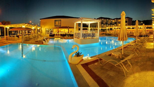 Une piscine avec des chaises longues et des parasols, un espace de détente couvert et un bâtiment doté de plusieurs balcons.