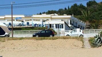 Un hotel con varios balcones, un primer plano con arena y coches aparcados.