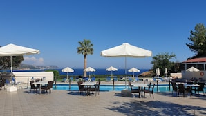 Zona junto a la piscina con mesas, sillas, sombrillas y vista al mar.