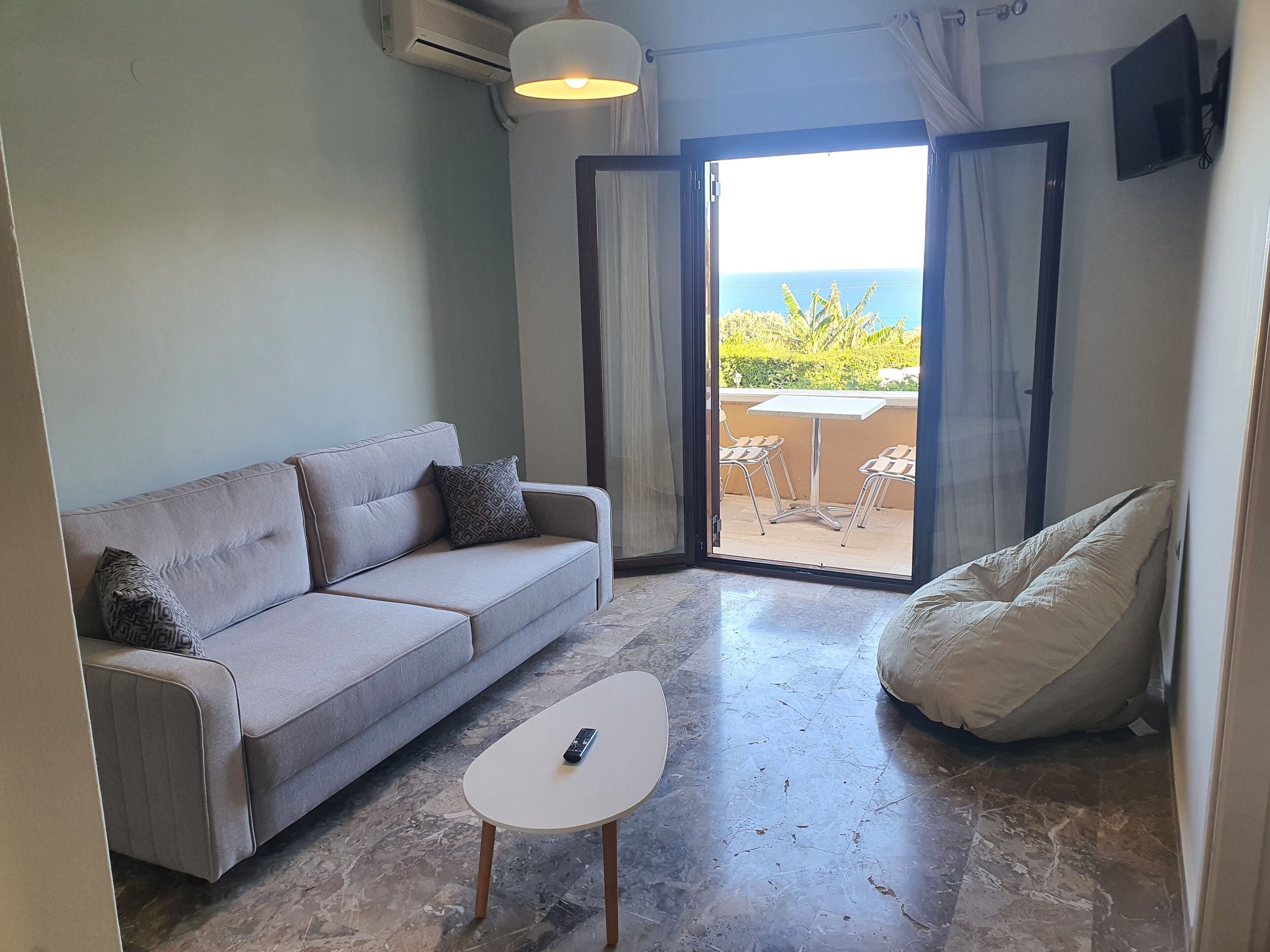 A modern living room with a grey sofa, a white coffee table, and a bean bag chair. A remote control is on the table. The room has a large sliding glass door leading to an outdoor area with a table and chairs.