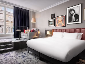 A hotel room with a large bed, a sofa, a desk, and a window with curtains. There are framed pictures on the wall.