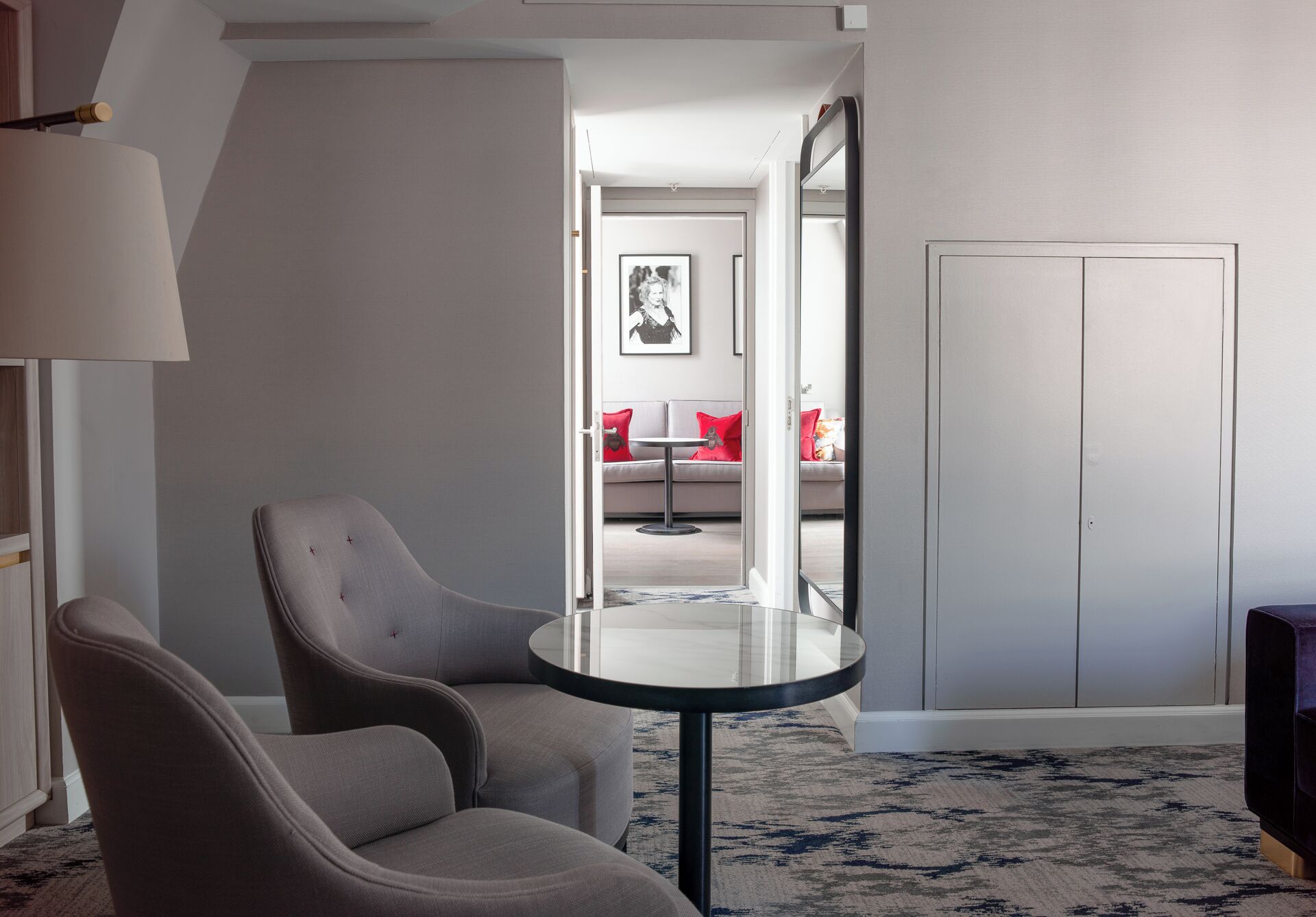 Una sala de estar moderna con una mesa redonda de vidrio, dos sillones grises, un sofá con cojines rojos y un armario.