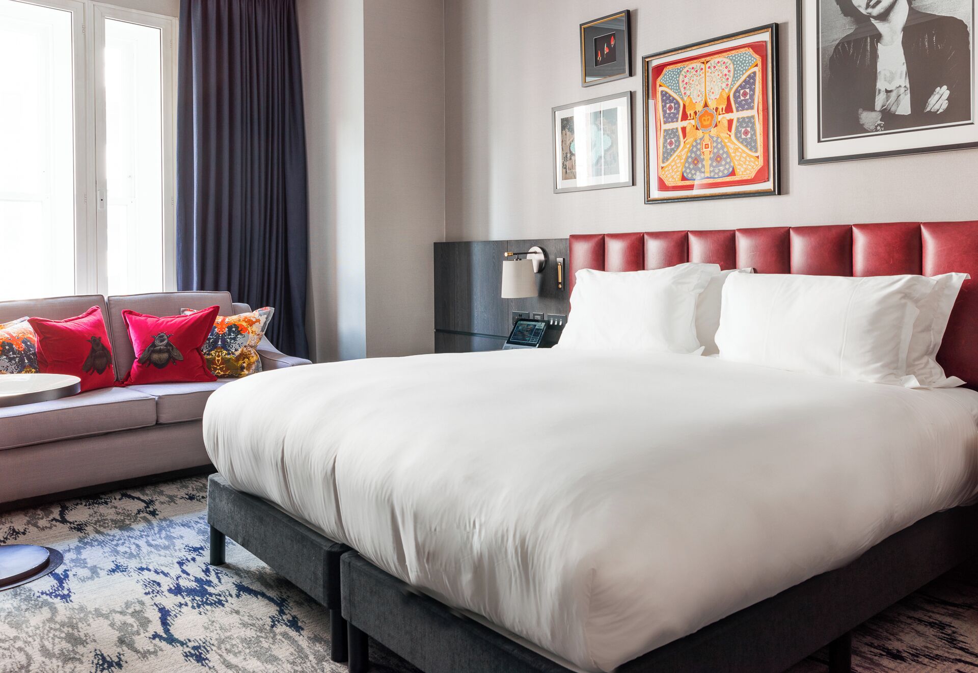 A hotel room with a large bed, a bedside table with a lamp, a sofa with colorful cushions, and artwork on the wall.