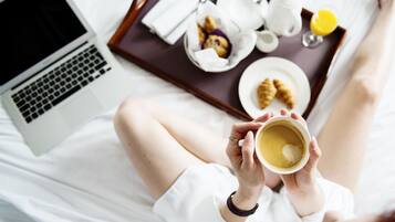 Uma pessoa segurando uma xícara de café, um laptop e uma bandeja com itens de café da manhã sobre a cama.