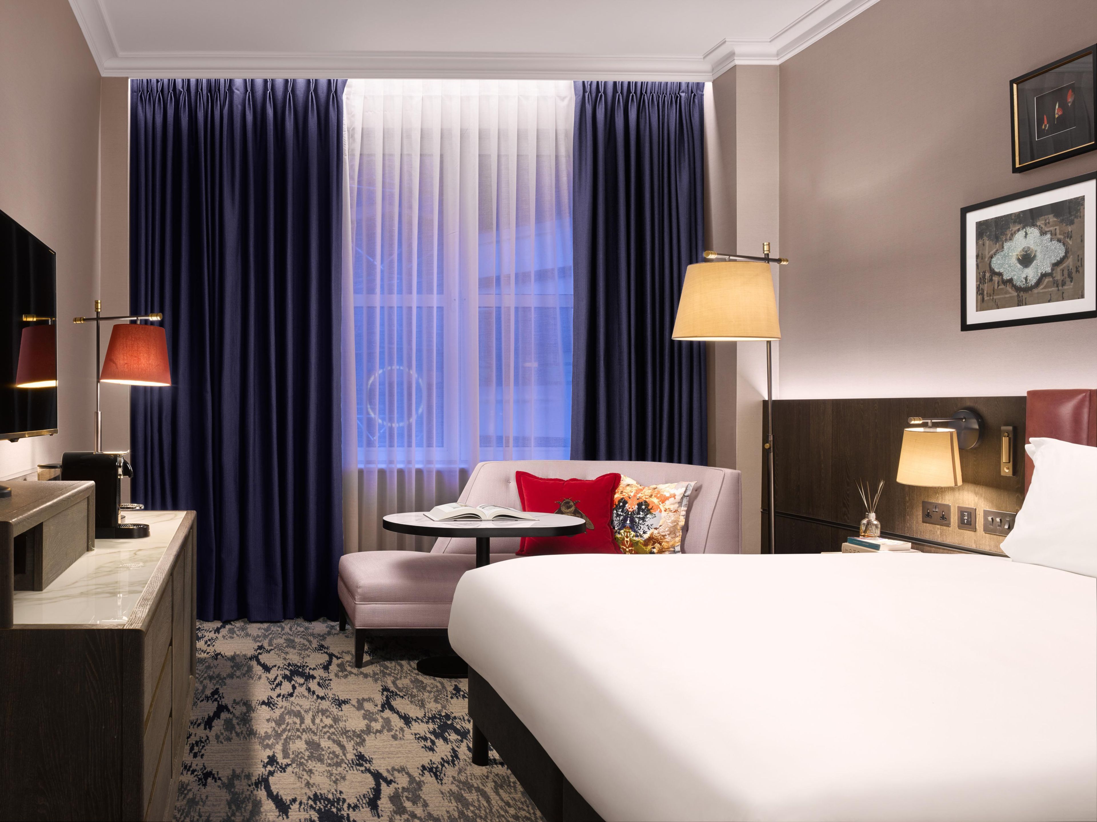 Ein Hotelzimmer mit einem Bett, einem Schreibtisch, einem Stuhl, einem kleinen Tisch mit einem Buch und einem Fenster mit Vorhängen.